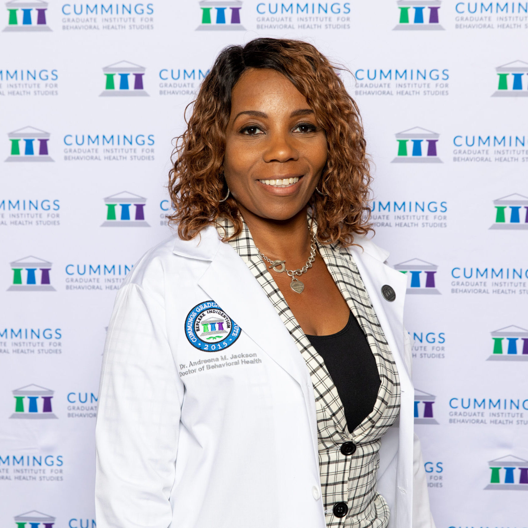 Dr. Andreena Jackson, Doctor of Behavioral Health (DBH) alumna, wearing a white coat in front of a Cummings Graduate Institute for Behavioral Health Studies backdrop.