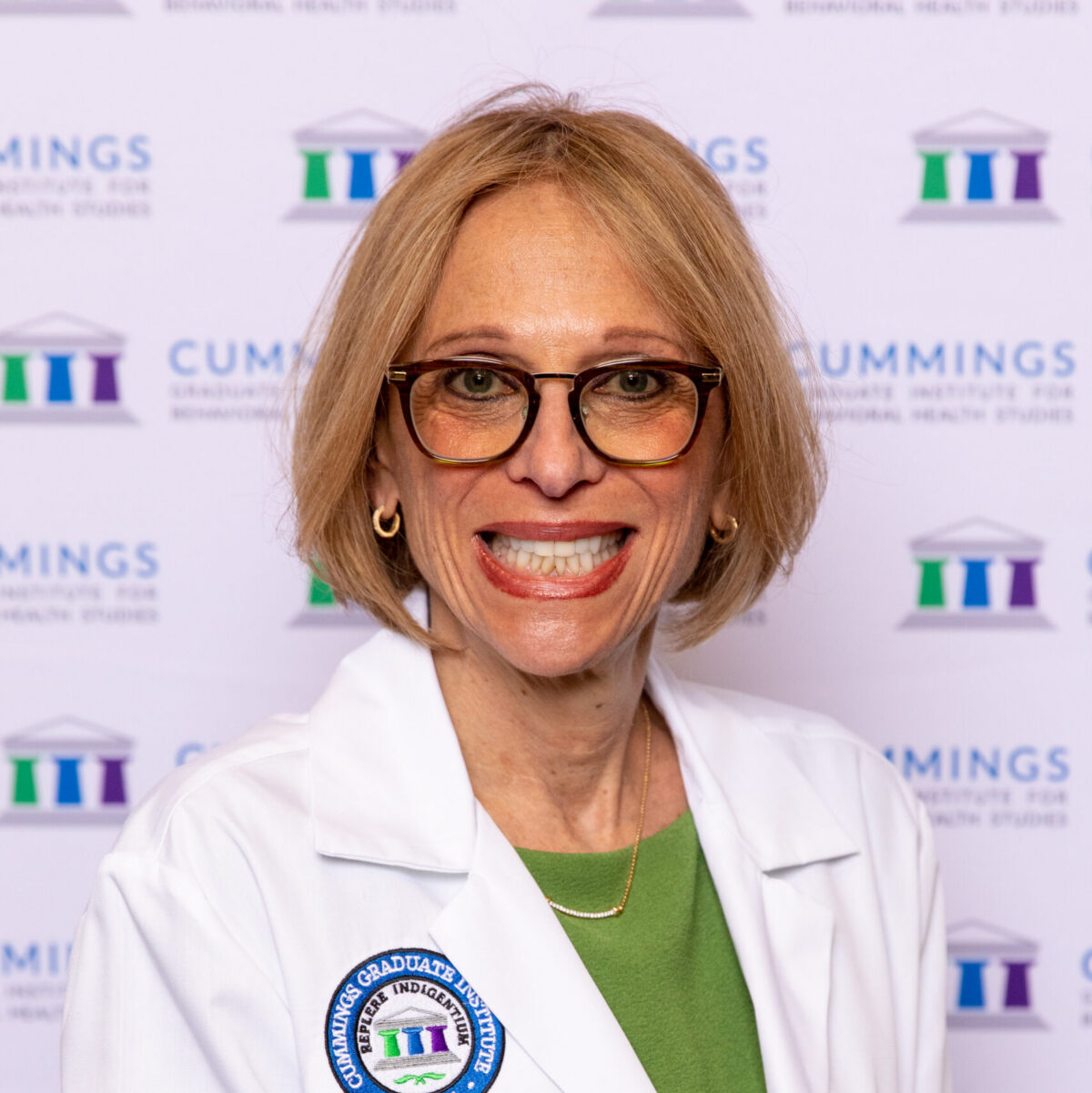 Dr. Ellen Fink-Samnick, , Doctor of Behavioral Health (DBH) alumna, wearing a white lab coat in front of a Cummings Graduate Institute for Behavioral Health Studies backdrop.