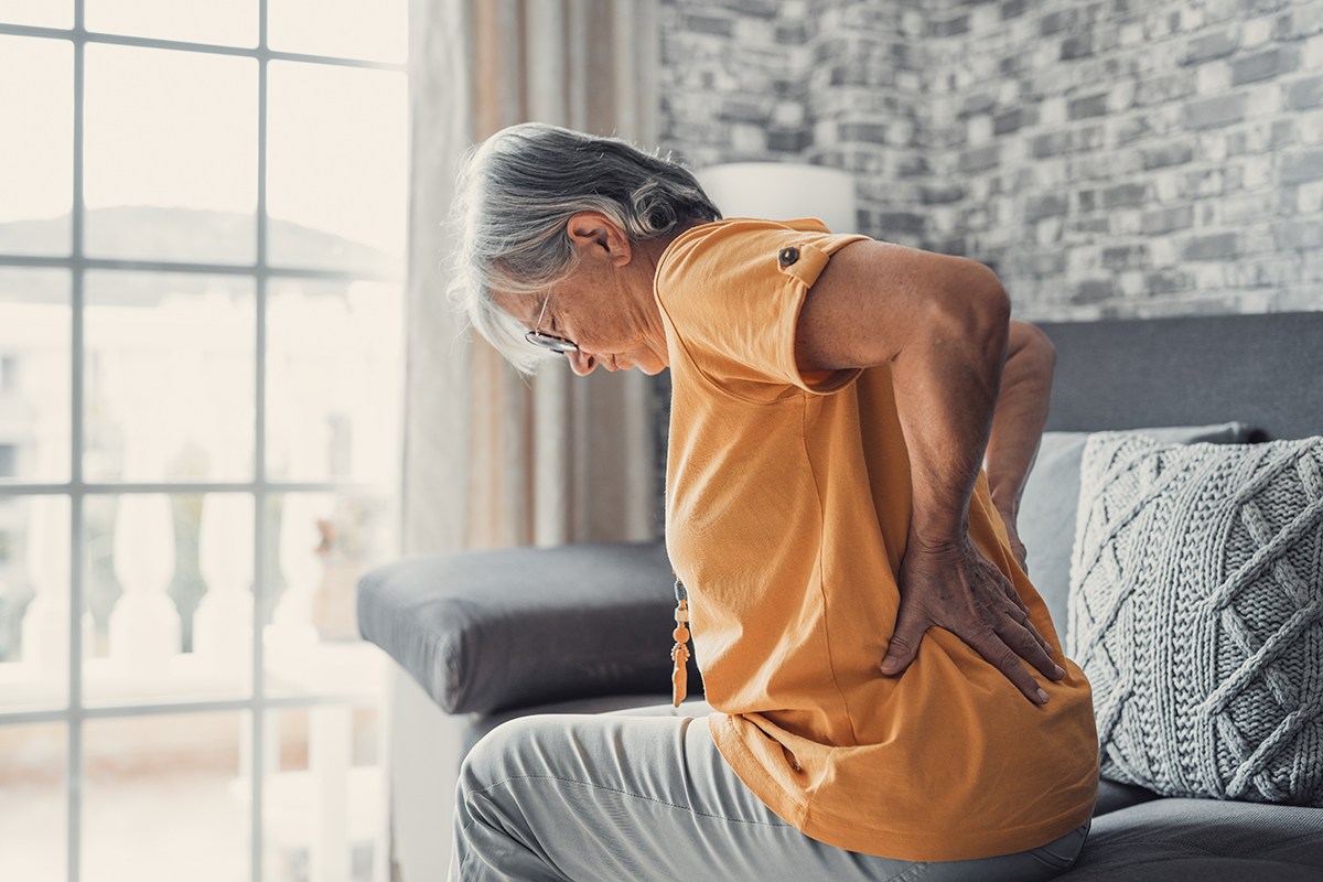 Unhappy mature woman touching back, feeling pain, sitting on couch in living room, unhealthy middle aged senior female suffering from kidney disease