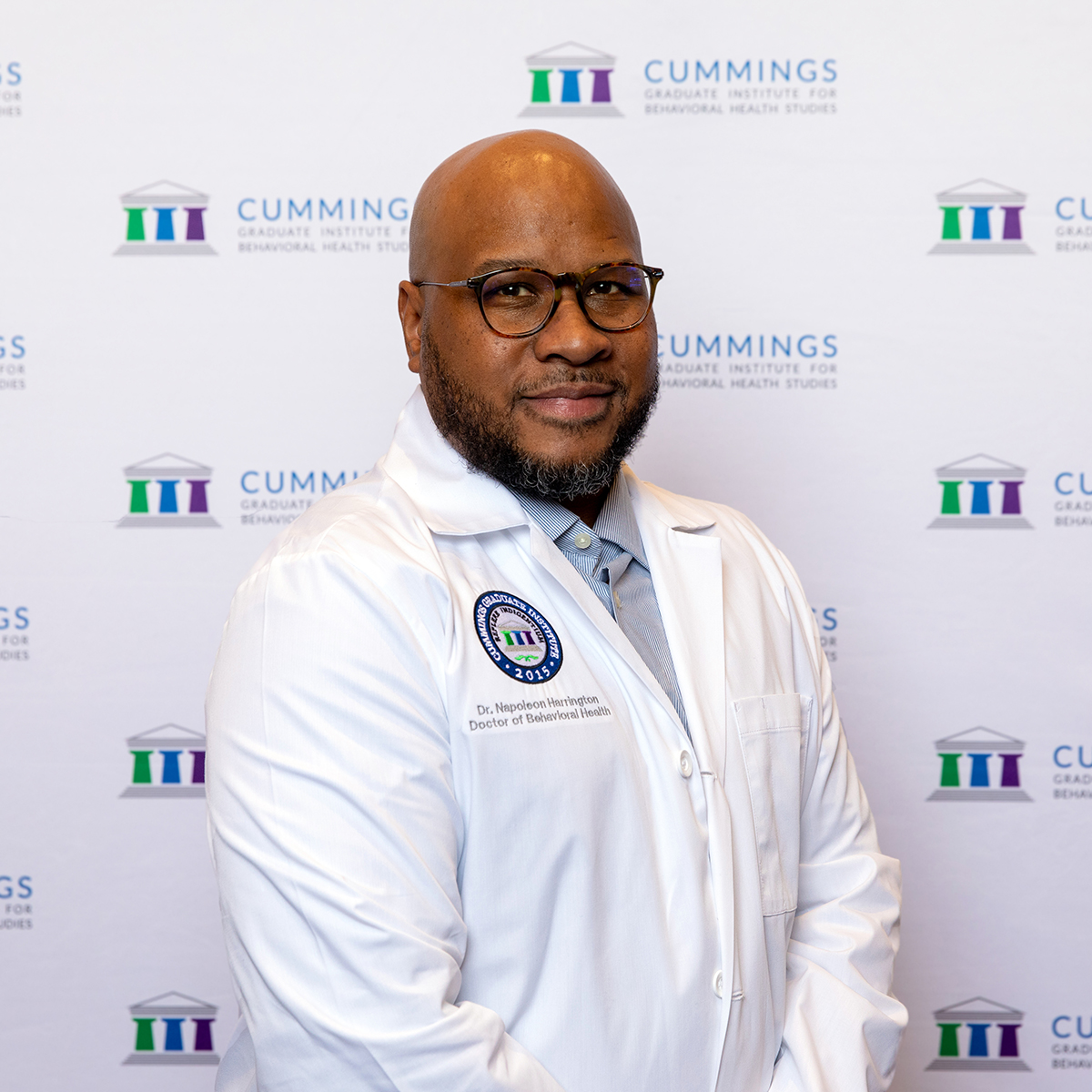Dr. Napoleon Harrington, Doctor of Behavioral Health (DBH) alumnus, wearing a white lab coat in front of a Cummings Graduate Institute for Behavioral Health Studies backdrop.