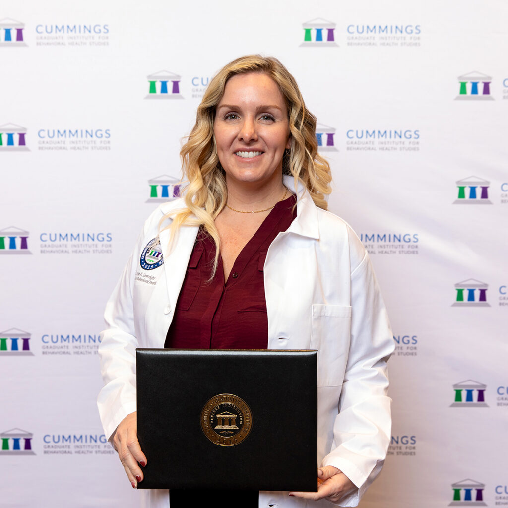 DBH graduate, Dr. Erin Enwright, wearing a white coat and holding a CGI diploma at commencement.