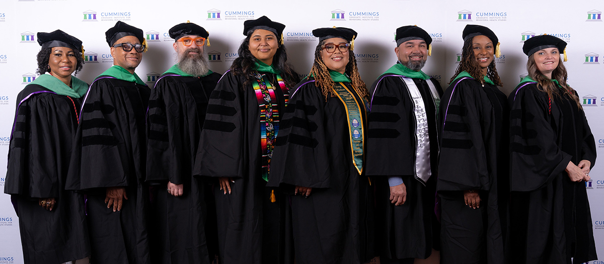 Eight Cummings Graduate Institute Doctor of Behavioral Health graduates stand together in academic regalia at the June 2026 commencement ceremony.