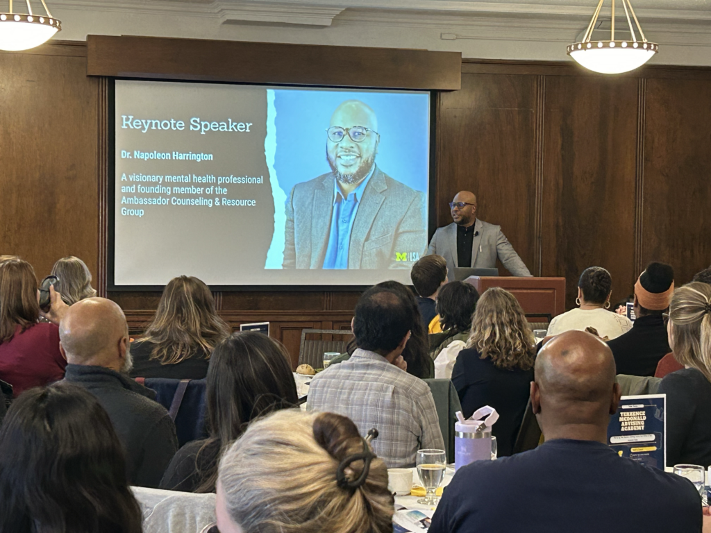 Dr. Napoleon Harrington delivering a keynote presentation to an audience, with a keynote speaker slide displayed behind him.