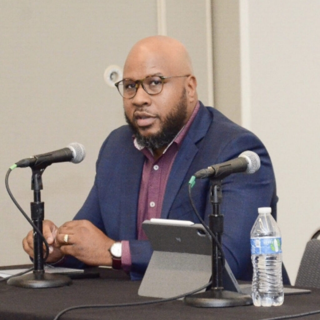 Dr. Napoleon Harrington speaking on a panel, seated at a table with microphones.
