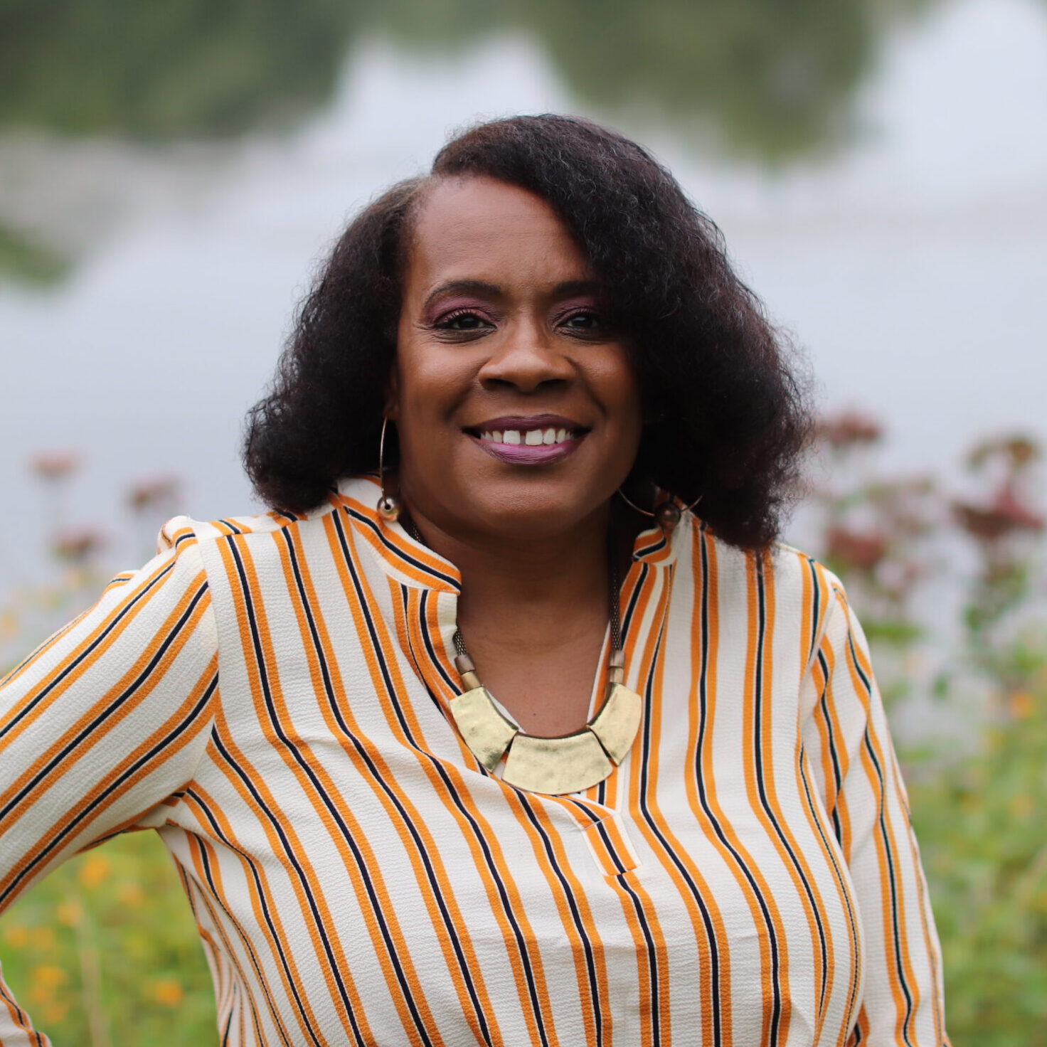 Portrait of CGI Doctor of Behavioral Health (DBH) student Lois Goudeau, M.Ed., LCPC, smiling outdoors near a lake, wearing a striped blouse and gold statement necklace.